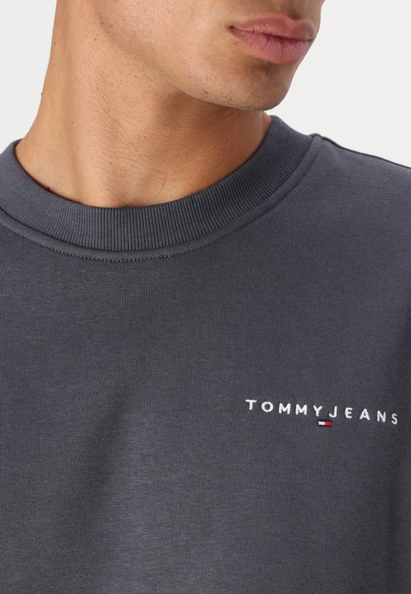 Felpa in cotone grigio scuro con collo a coste e logo ricamato "TOMMYJEANS" in bianco. Presenta piccoli accenti rossi e blu.