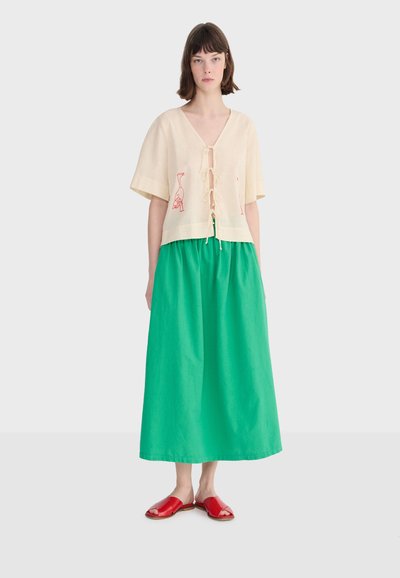 Femme portant un haut crème noué sur le devant avec une broderie d'oiseau rouge, une longue jupe verte et des sandales ouvertes rouges sur fond blanc.