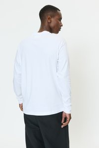 Vit långärmad t-shirt i bomull med rund halsringning och rak fåll, bärs tillsammans med svarta byxor.