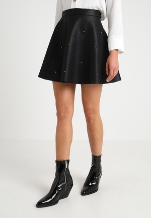 Vrouw die een zwarte uitlopende rok met kleine studs draagt, een wit shirt met lange mouwen en glanzende zwarte enkellaarsjes met sleehak, staand op een witte vloer.