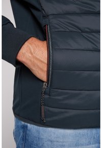 Jachetă quilted de culoare bleumarin cu buzunare laterale cu fermoar, manșete elastice și un material texturat. Prezintă o etichetă cu logo în apropierea fermoarului.