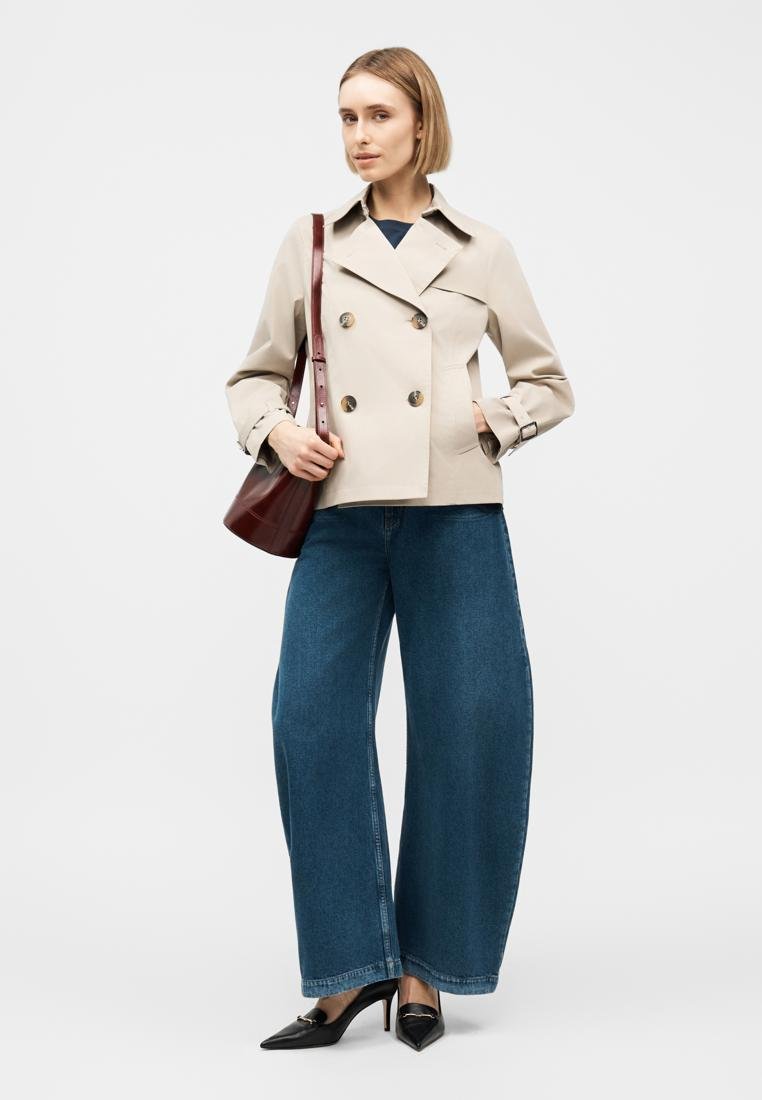Femme portant une veste beige à double boutonnage, un jean bleu à jambes larges, des talons noirs pointus, et un sac à bandoulière rouge foncé, sur fond uni.