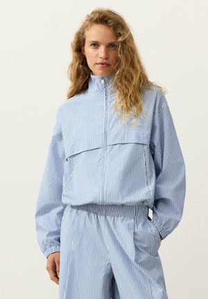 Donna con lunghi capelli ricci che indossa una giacca con zip a righe verticali blu e bianche e pantaloni coordinati, in piedi con le mani nelle tasche.