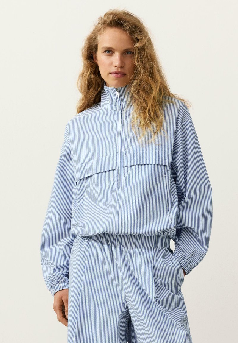 Femme aux cheveux longs et bouclés portant une veste zippée à rayures verticales bleues et blanches et un pantalon assorti, debout, les mains dans les poches.