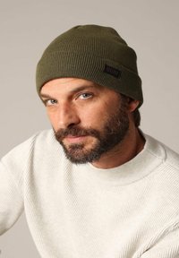 Bonnet tricoté vert olive avec une texture côtelée et une petite étiquette noire portant l'inscription "DEELUXE". Porté avec un pull de couleur claire.