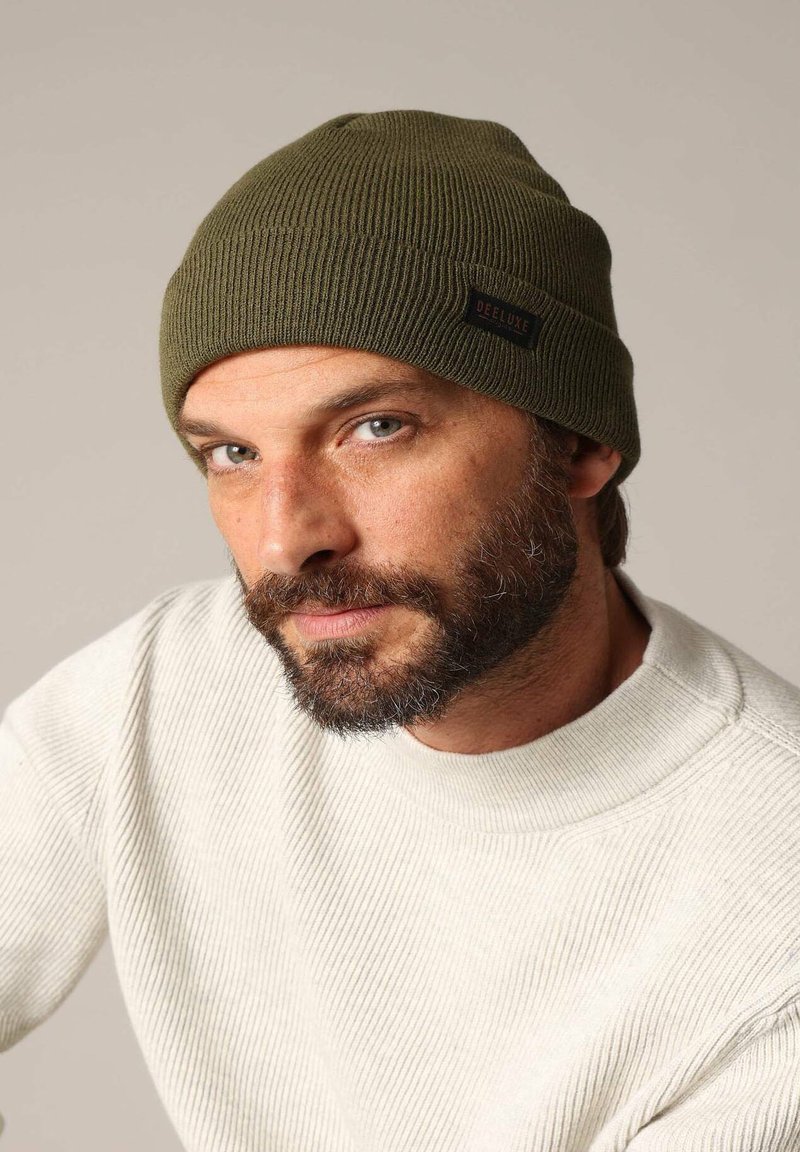 Bonnet tricoté vert olive avec une texture côtelée et une petite étiquette noire portant l'inscription "DEELUXE". Porté avec un pull de couleur claire.