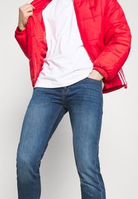Red puffer jacket with quilted texture, white T-shirt underneath, and dark blue slim-fit denim jeans with a slight stretch.