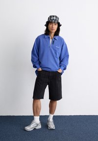 Sweatshirt azul de fleece com colarinho de zíper, calções pretos às riscas, sapatilhas prateadas e um chapéu de balde preto e branco com padrão xadrez.
