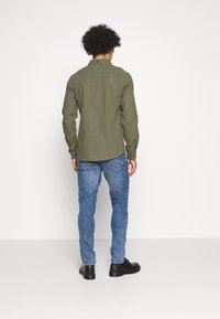 Olivově zelená košile s dlouhým rukávem a límečkem, v kombinaci s modrými džíny a černými botami. Košile má rovný střih a texturovanou látku.