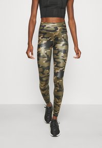 Legginsy w kamuflażu w odcieniach zieleni i czerni, wykonane z błyszczącego, elastycznego materiału; zestawione z czarnym crop topem i czarnymi butami sportowymi.