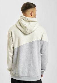 Hooded sweatshirt with a two-tone design: cream upper section with flecks, gray lower section. Soft fabric, relaxed fit, visible stitching.