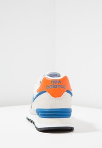 Baskets en suède blanc et mesh avec des accents orange et bleu, présentant un talon texturé et un logo "new balance" à l'arrière. Semelle extérieure noire.