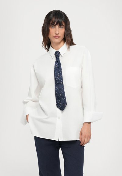 Chemise blanche à manches longues avec une poche poitrine. Assortie à une cravate bleu marine à petit motif. Coupe décontractée avec les manches retroussées.