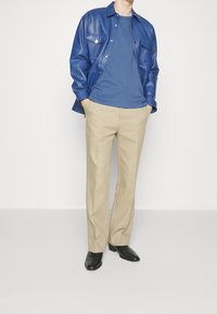 Blauwe neplederen shirtjacket, blauwe rondhals t-shirt, beige nette broek, zwarte schoenen. Details omvatten voorzakken en knoopsluitingen.