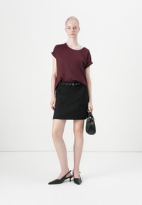 Burgunderröd kortärmad skjorta ihop med en svart mini kjol med bälte i midjan. Modellen håller en liten svart handväska och bär svarta slingback-klackar.