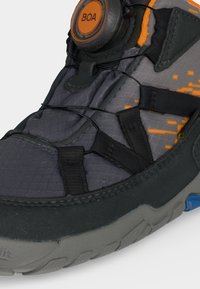 Gros plan d’une chaussure grise et noire avec des détails orange, équipée d’un système de fermeture BOA à cadran et de lacets fins noirs sur une tige en tissu texturé.