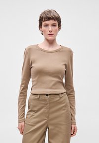 �Μάκρυ μανίκι, ανοιχτό καφέ μπλουζίτσα με στρογγυλή λαιμόκοψη, φτιαγμένη από μαλακό ύφασμα. Συνδυασμένη με ανοιχτό καφέ παντελόνια που διαθέτουν μπροστινή τσέπη.