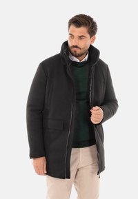 Cappotto di lana nera con chiusura a zip, colletto alto e due tasche frontali. Indossato sopra un maglione verde scuro e pantaloni beige chiaro. Design semplice.