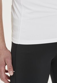 Vit snabbtorkande träningsskjorta med mesh-sidor, tillsammans med svarta åtsittande leggings. Handen vilar på leggings vid sömmen.