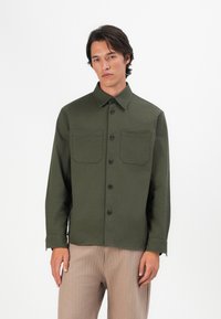 Joven vistiendo una camisa verde oliva de botones con dos bolsillos en el pecho y pantalones beige, de pie frente a un fondo blanco liso.
