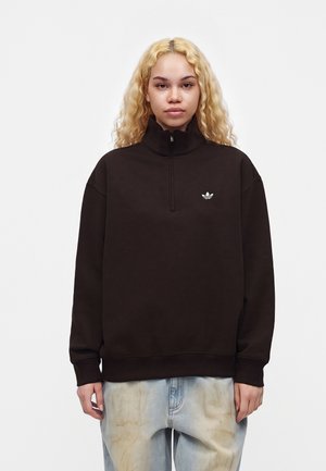 Jong persoon met lang blond krullend haar, gekleed in een zwarte trui met een kwart ritssluiting, met een klein wit Adidas-logo en lichtgewassen spijkerbroek.
