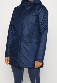 Marineblauwe geïsoleerde parka met een rits en zakken. Heeft een capuchon en een gladde, glanzende stofstructuur. Geschikt voor koud weer.