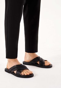 Sandale negre din piele, cu barete încrucișate, talpă plată și vârf deschis, asociate cu pantaloni negri eleganți.