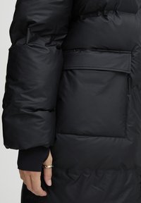 Cappotto imbottito nero con un'esterno liscio e lucido, maniche testurizzate e una tasca orizzontale su un lato. I polsini elastici completano il design.