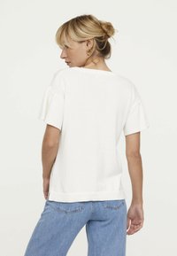 Pull en tricot blanc avec des manches courtes évasées et un ourlet côtelé. Associé à un jean en denim bleu clair, affichant une coupe décontractée.