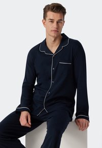 Tummansininen pyjamapaita pehmeästä kankaasta, jossa valkoiset reunat, sisältäen nappikiinnitteisen paidan ja pitkät housut, sekä taskun vasemmassa rinnassa.