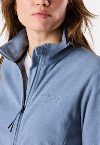 Giacca in pile blu chiaro con zip frontale, colletto alto e logo ricamato. Il tessuto sembra morbido con una finitura leggermente texturizzata.