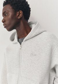 Jeune homme aux cheveux courts et bouclés, portant un sweat à capuche gris clair zippé sur un t-shirt rayé, regardant vers la gauche sur un fond uni.