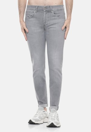 Persona che indossa jeans grigi chiari slim-fit con risvolti e scarpe da ginnastica bianche, in piedi davanti a uno sfondo semplice.