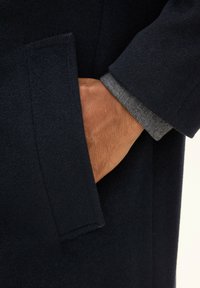 Marine uldfrakke med en sidelomme og en synlig skjorteærme i grå og hvid mønster. Stoffet har en glat tekstur.