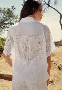Camicia bianca in cotone con intricati tagli a foro, colletto, maniche corte e vestibilità ampia. Mostrata di spalle.