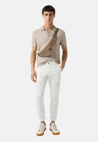 Beige skuldertaske med logo, båret over brystet, parret med lys beige polo-shirt, hvide bukser og hvide sneakers med brune såler.