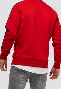 Uomo che indossa una felpa rossa con strisce blu sulle maniche, una maglietta bianca sotto, jeans grigi e un anello nero sulla mano destra.