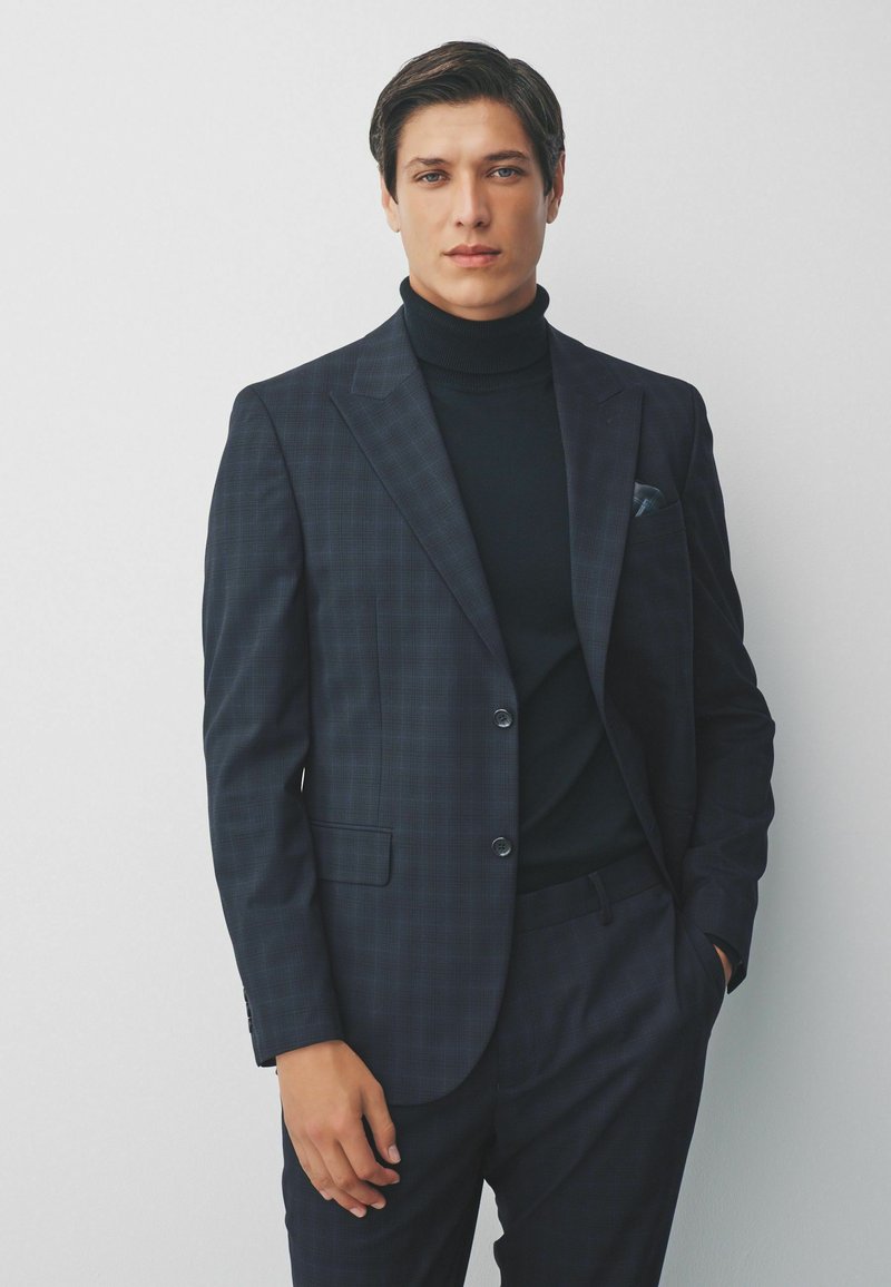 Marineblauw geruite pak met een slim fit, enkele knoopsluiting en twee voorzakken, gecombineerd met een zwarte coltrui en een bijpassend pochet.