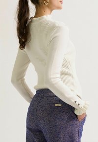 Vrouw met een hoge paardenstaart, die een witte geribbelde top met lange mouwen en knoopmanchetten draagt, samen met blauwe textuur hoge taille broek, met haar hand in de zak.