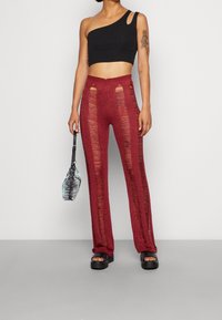 Žena v černém topu na jedno rameno, červených pletených kalhotách s vertikálními roztrženými pruhy, černých platformových sandálech a držící malou kabelku se zebřími pruhy.