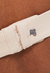 Beige elastisk rem med et lukningspunkt og grå broderet detalje, sat mod en brun tekstureret baggrund.