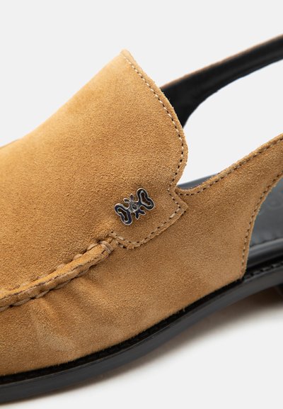 Ταν δερμάτινο παπούτσι τύπου slip-on με μαύρη σόλα, λεπτομερείς ραφές και μικρή μεταλλική διακόσμηση λογότυπου κοντά στην πάνω άκρη.