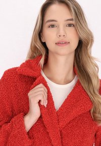 Czerwona, teksturowana polarowa kurtka z szerokim kołnierzem, z guzikiem z przodu i kieszeniami. Modelka nosi pod spodem kremową bluzkę.