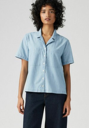 Camisa de manga corta azul claro en tejido suave, con botones delanteros, cuello y dos bolsillos en el pecho. Diseño sencillo con un ajuste relajado.