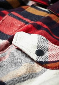 Rutet flannel stof med rød, marineblå, grå og hvide farver. Udsmykket med en sort trykknap og en struktureret, blød finish.