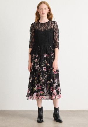 Sort lacekjole med lange ærmer og peplum-talje, der har et blomsterprint på en sort nederdel. Sorte ankelstøvler fuldender looket.