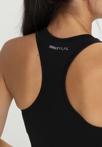 Pogled od zadaj na žensko, ki nosi črno športno majico z zadajšnjim pasom v slogu dirkača, na kateri je blizu roba vratu natisnjen logotip "ONLY PLAY".