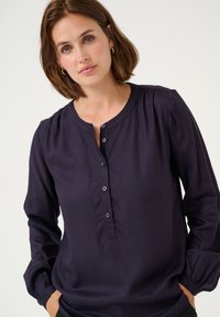 Blouse bleu marine à manches longues avec un col rond, patte de boutonnage et manches froncées. Texture lisse, tissu doux.