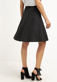 Jupe évasée noire avec une texture lisse, de conception mi-longue, portée avec des sandales à talons noires présentant des découpes et des détails à lacets.