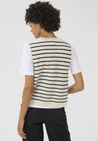 Una donna con capelli ricci indossa un gilet a strisce sopra una camicia bianca, girata di spalle, mostrando il motivo a strisce nere e crema.
